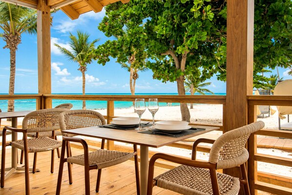 An outdoor dining area with a wooden table and wicker chairs, overlooking a beach with palm trees and the ocean.