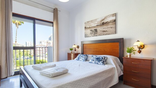 A hotel room with a large bed, a wooden headboard, two bedside tables with lamps, a view of the city through a balcony, and a beach scene painting on the wall. - B97 - Flor da Rocha Apartment in Portim o (Portimão)