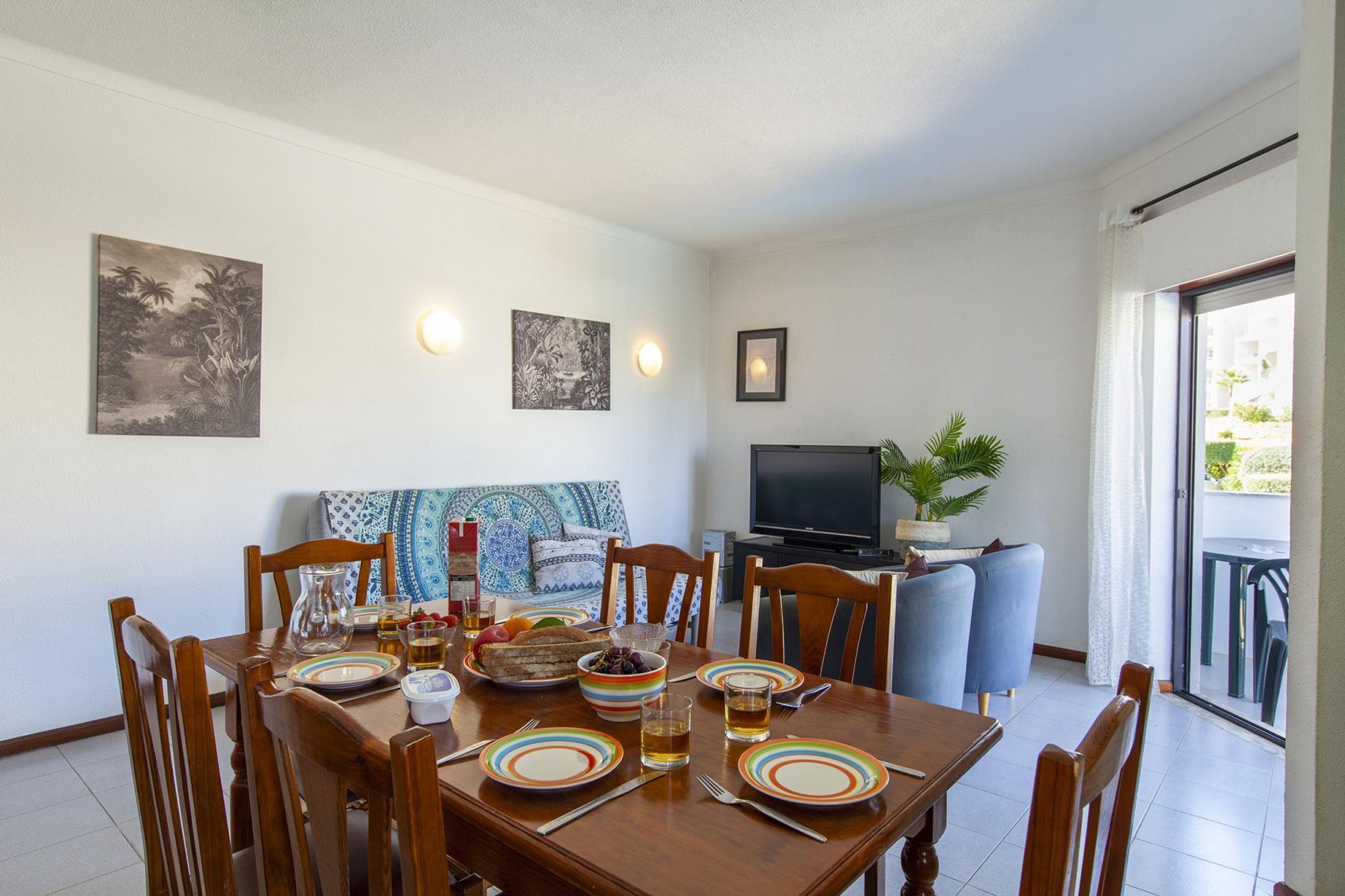 A dining area with a wooden table set for four, surrounded by chairs. A television is mounted on the wall, and there is a potted plant on a small table.