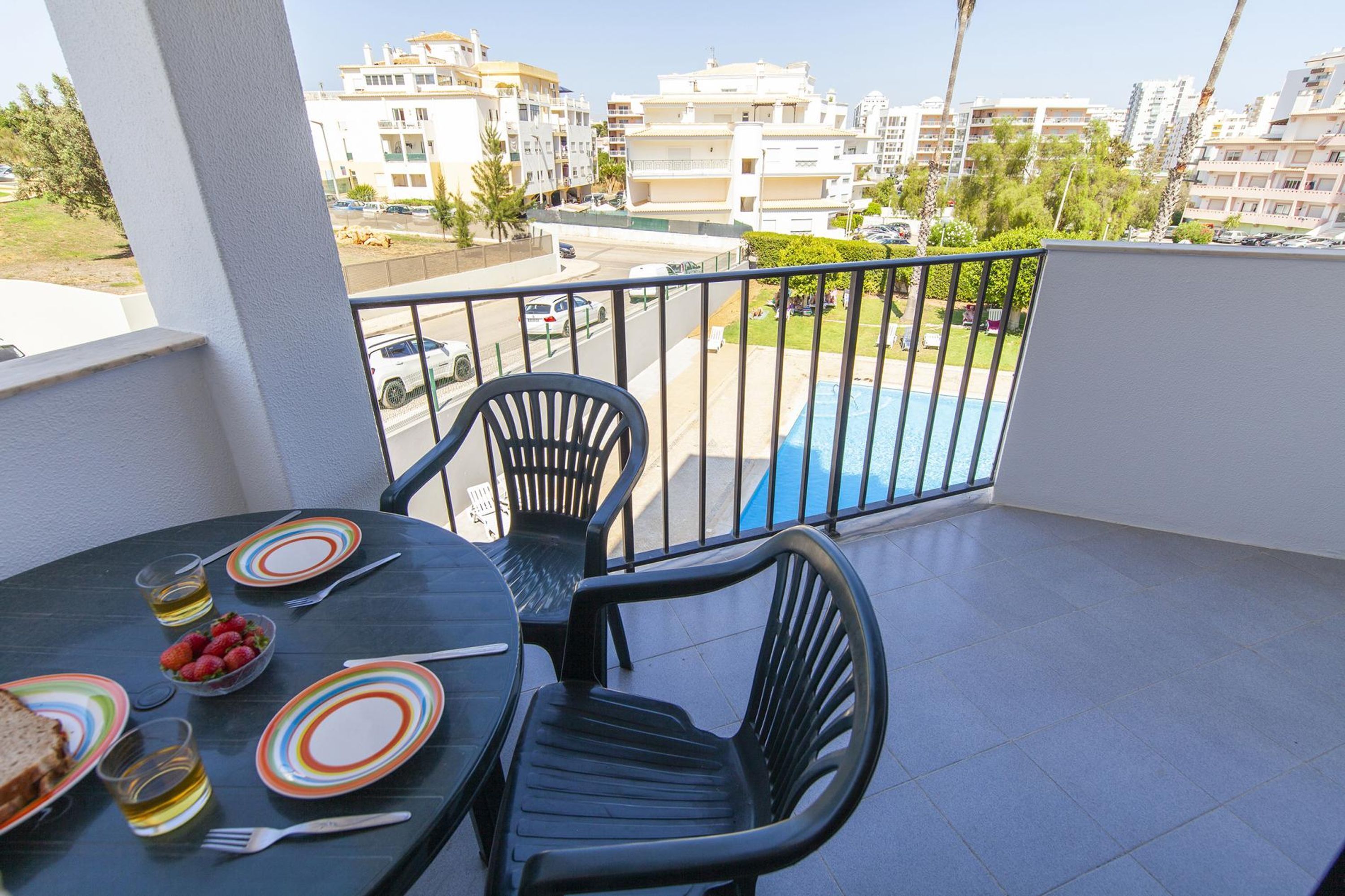 A balcony with a table set for two, chairs, and a view of a swimming pool and buildings in the background.