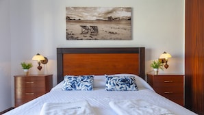 A neatly made bed with two pillows, two bedside tables with lamps, and a painting of a beach scene above the bed. - B97 - Flor da Rocha Apartment in Portim o (Portimão)