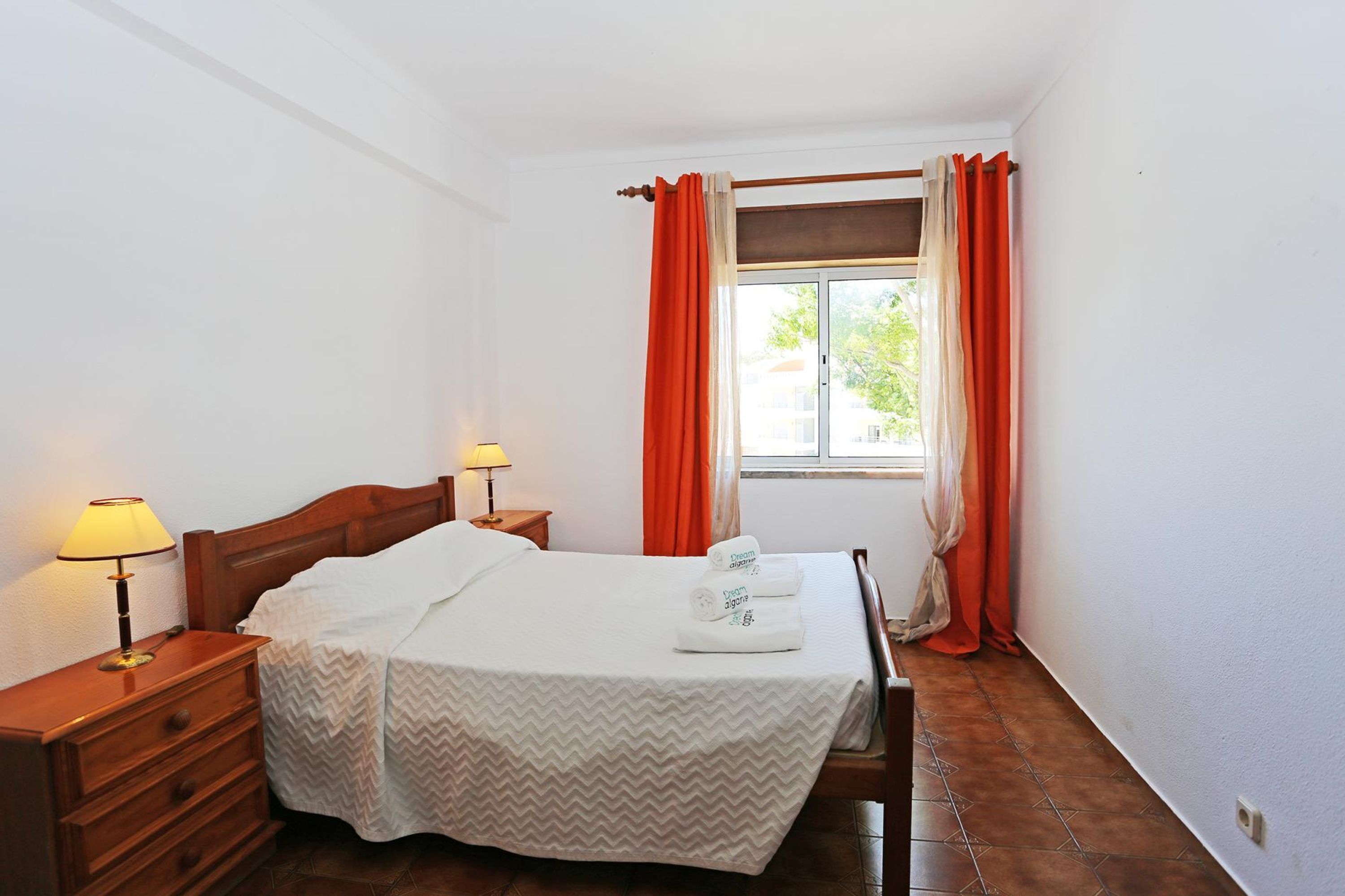 A neatly made bed with a white bedspread, two bedside lamps, and a window with orange curtains.