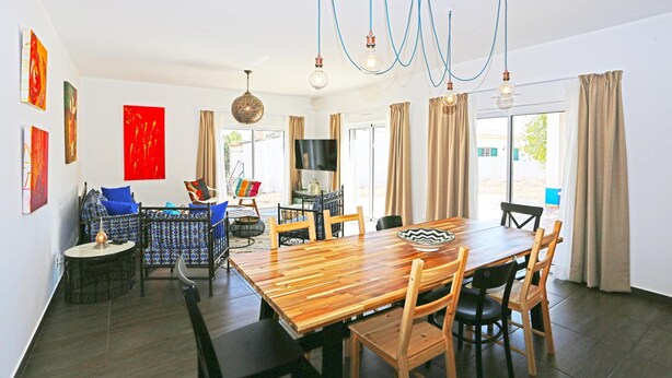Un área de comedor con una mesa de madera, sillas y una araña. Hay cortinas en las ventanas y obras de arte en las paredes.