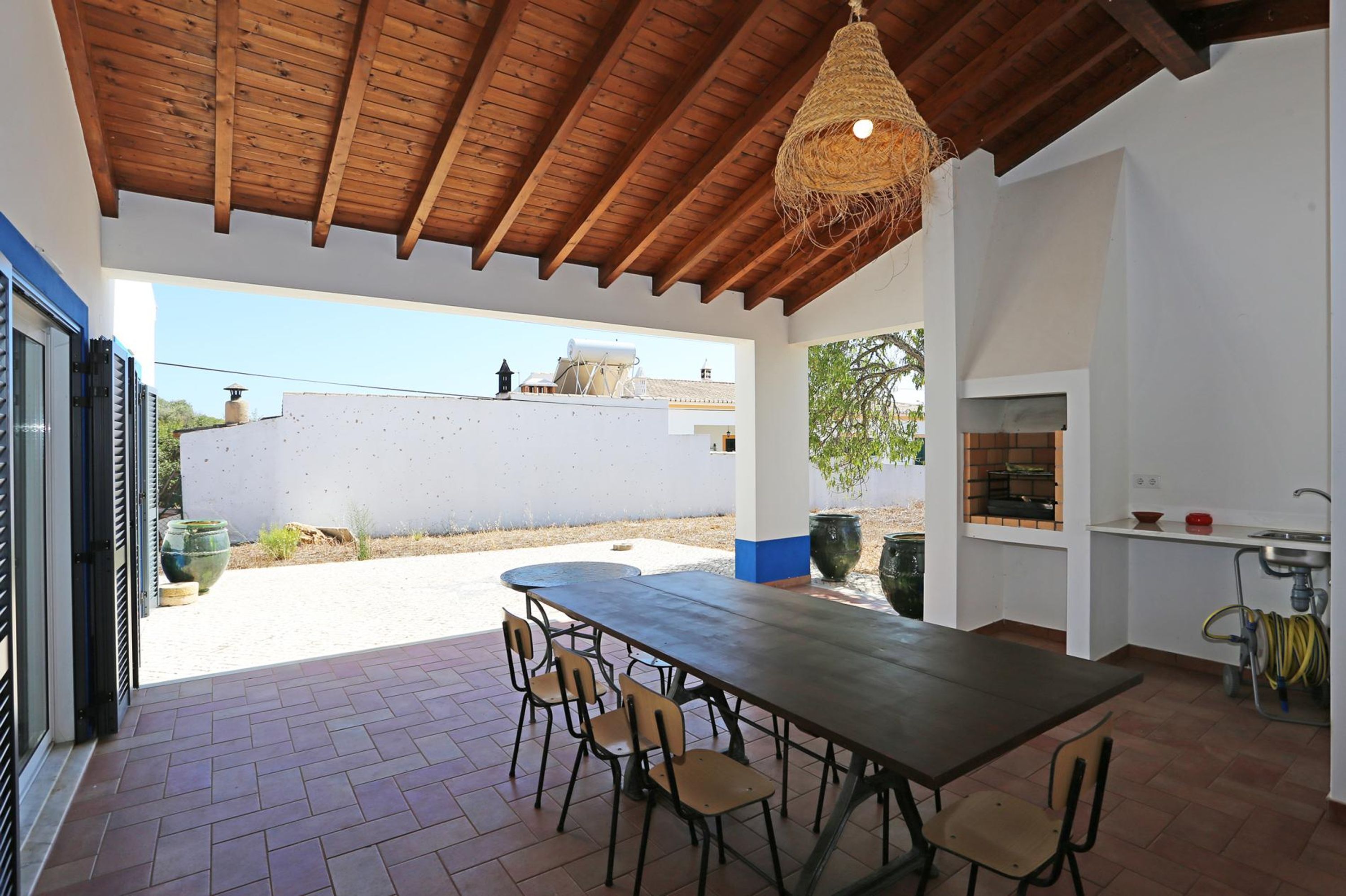 An outdoor patio with a wooden ceiling, a dining table with chairs, a hanging light fixture, and a kitchen area with a sink and a brick oven.