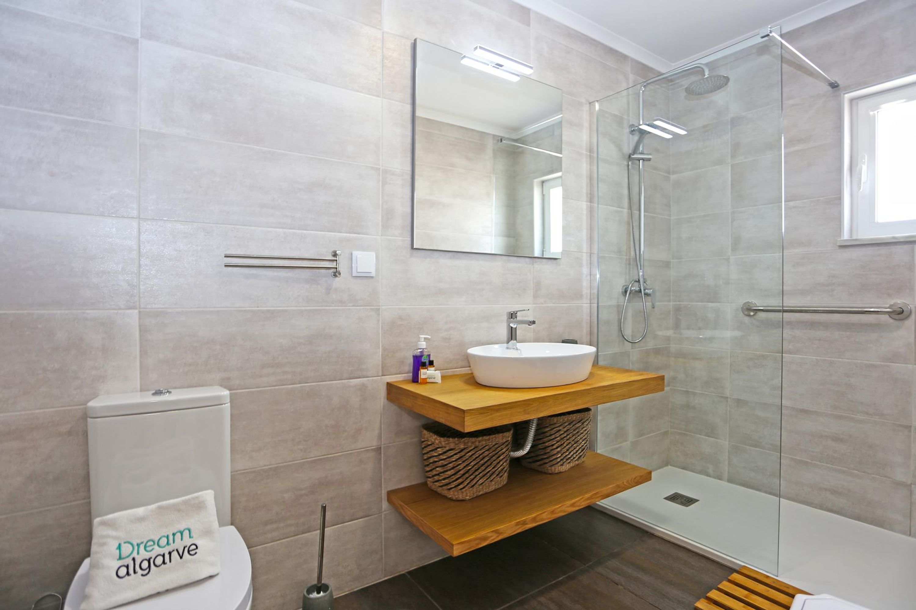A modern bathroom with a glass shower enclosure, a wooden vanity, and a toilet with a towel reading "Dream algarve".