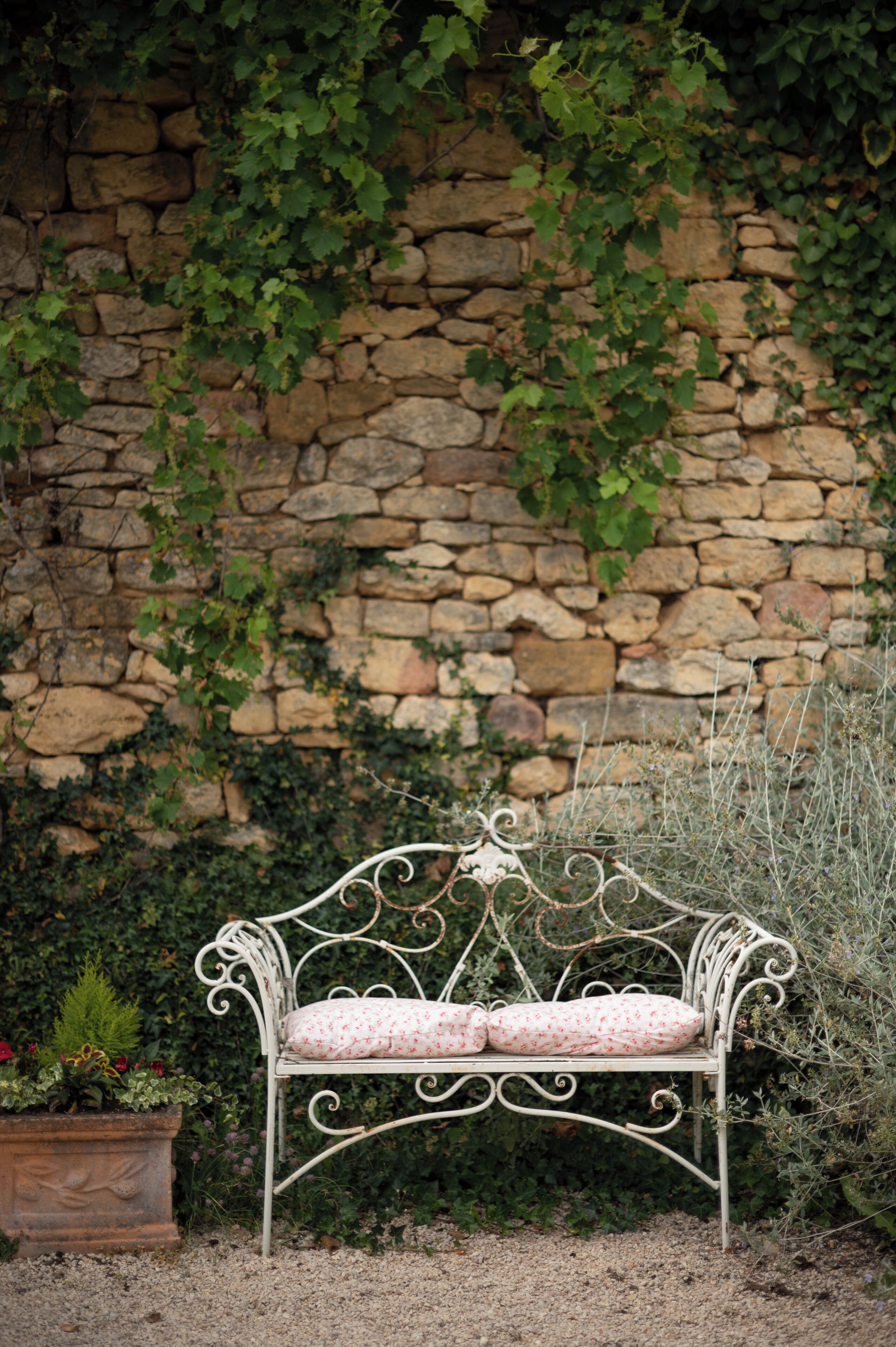 A wrought iron bench with a floral cushion, positioned in front of a stone wall with climbing plants.