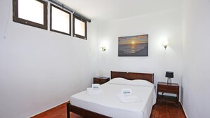 A bedroom with a bed, two bedside tables, a nightstand, a painting of a sunset, and a window with blinds.