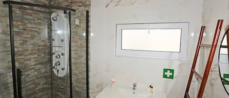Une salle de bain avec une douche en verre, un lavabo blanc avec un miroir au-dessus, et un symbole vert de premiers secours sur le mur.