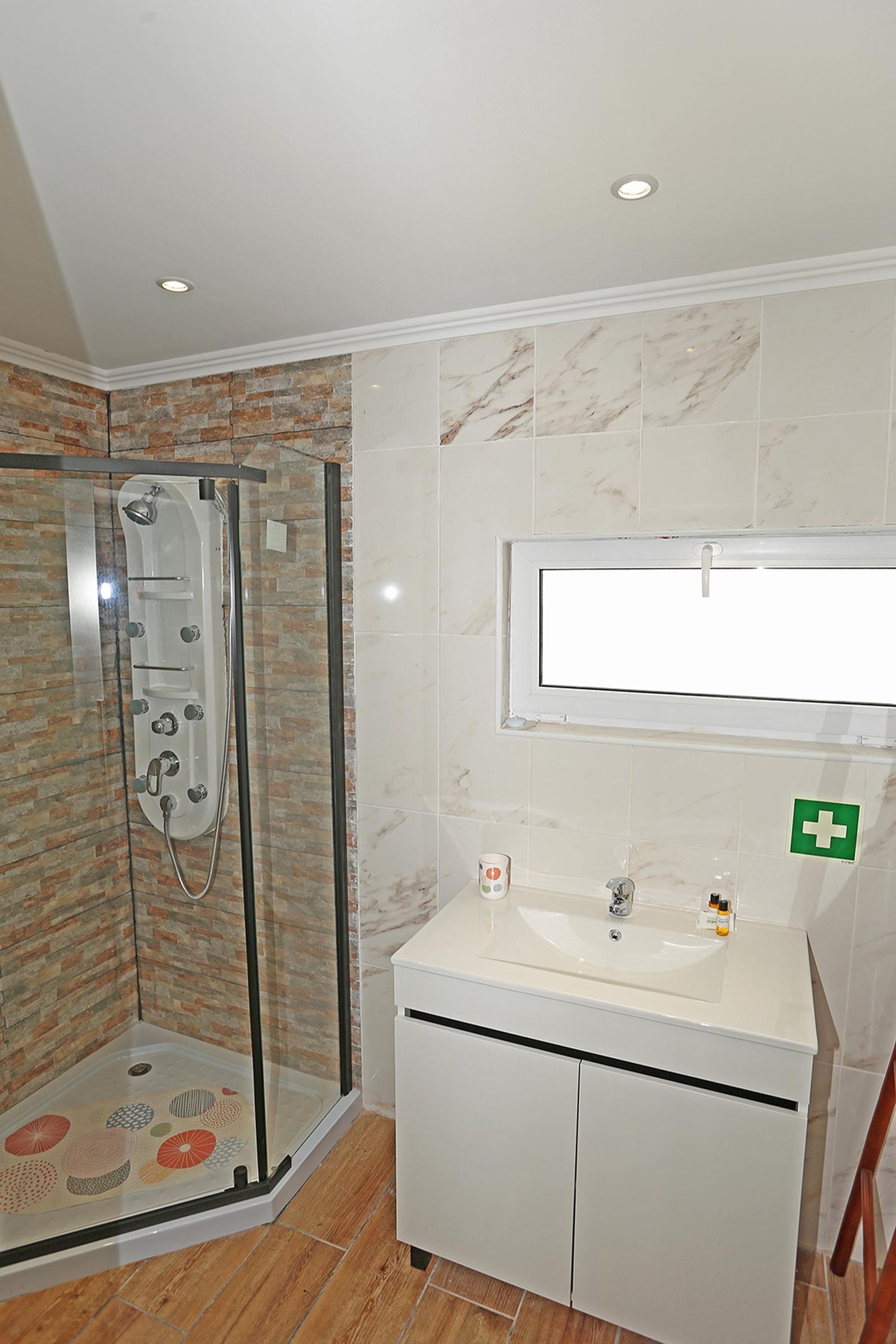 Une salle de bain avec une douche à parois de verre, un lavabo blanc avec un miroir au-dessus, et un symbole de premiers secours sur le mur.