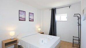A hotel room with a bed, two bedside lamps, a window with a curtain, and a wall-mounted rack. - X 06 - 1 Bed Apartment in Quinta da Ponte (Lagos)