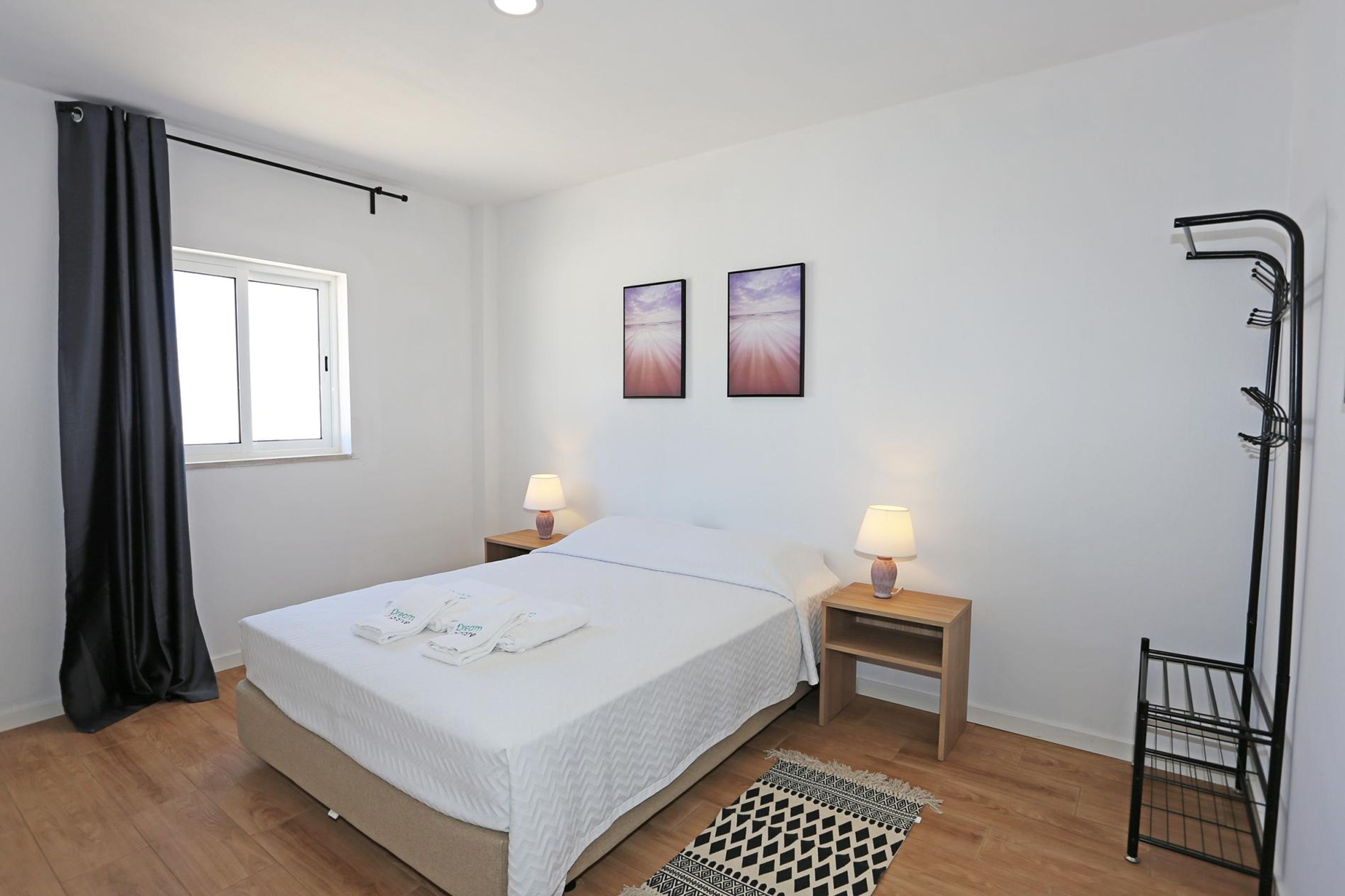 Ein ordentlich eingerichtetes Schlafzimmer mit einem Bett, Nachttischlampen, einem Fenster mit Vorhängen und zwei an der Wand hängenden Bildern.