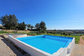 A rectangular swimming pool with a white border, surrounded by a paved area and greenery.
