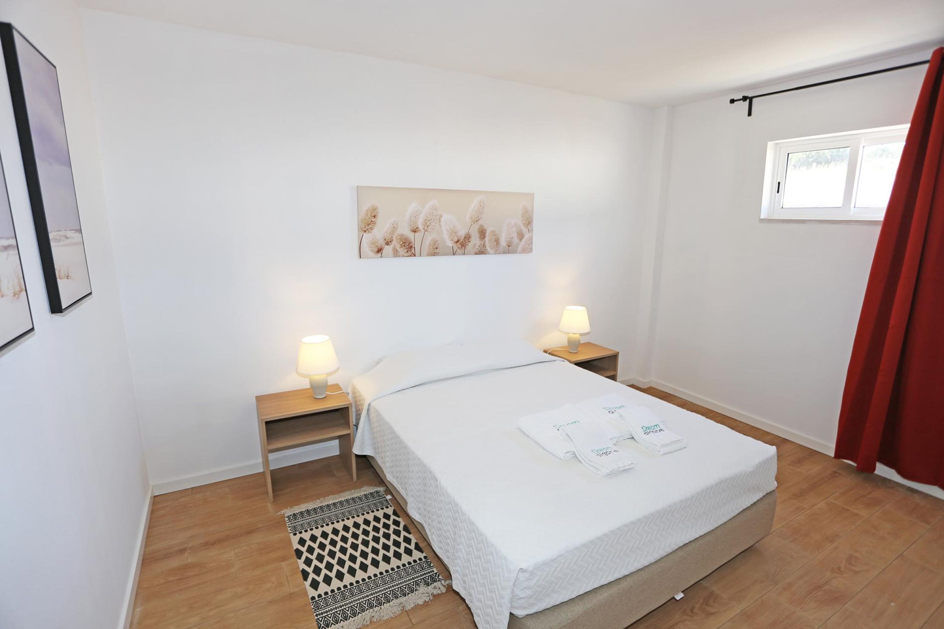Un dormitorio con una cama, dos mesitas de noche con lámparas, una alfombra, una ventana con cortinas y un cuadro en la pared.