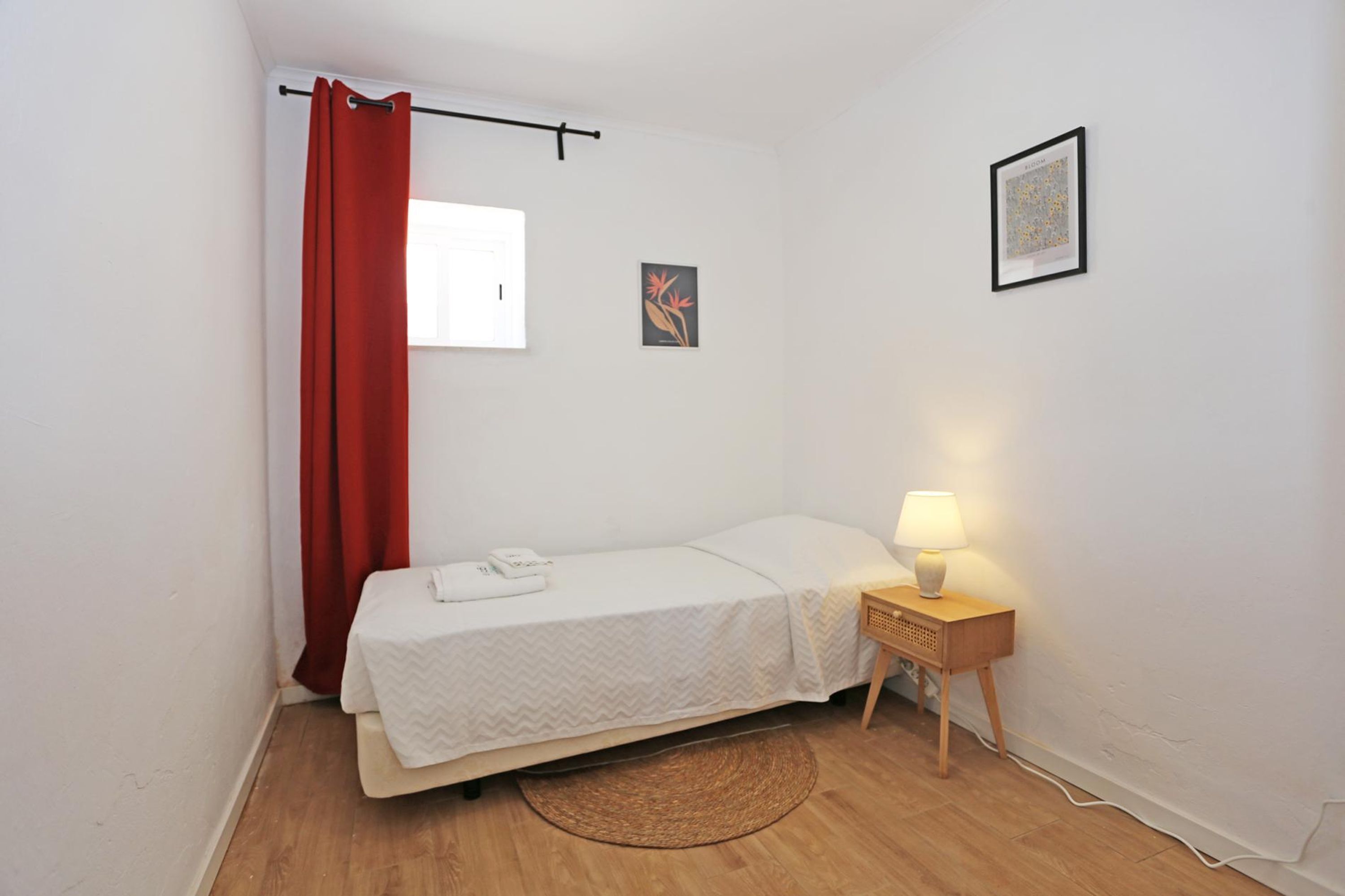 Een kleine, keurig opgeruimde slaapkamer met een eenpersoonsbed, een nachtkastje met een lamp, een rood gordijn en twee ingelijste foto’s aan de muur.