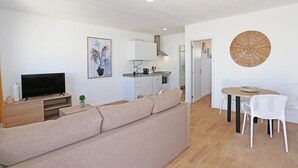 Een moderne woonkamer met een bank, televisie en een kleine plant. Er is een keukenhoek met kasten en een fornuis. Ook een eettafel met stoelen is te zien.