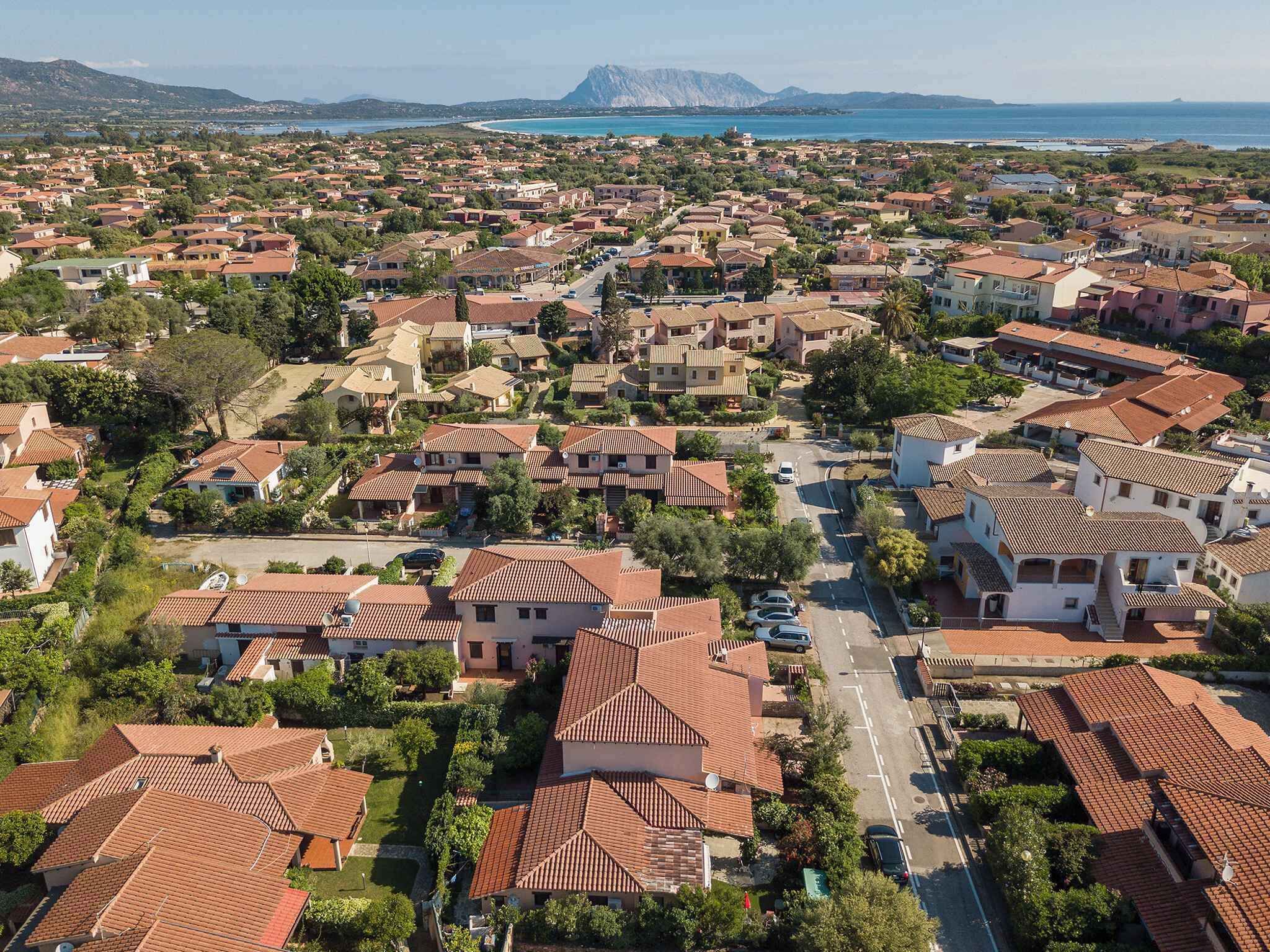 An aerial view of a residential area with terracotta-roofed houses, a main road, and a coastline in the background.