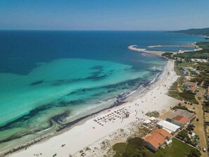 An aerial view of a beach with clear turquoise waters, a sandy shore, and a small coastal town with buildings and parked cars.