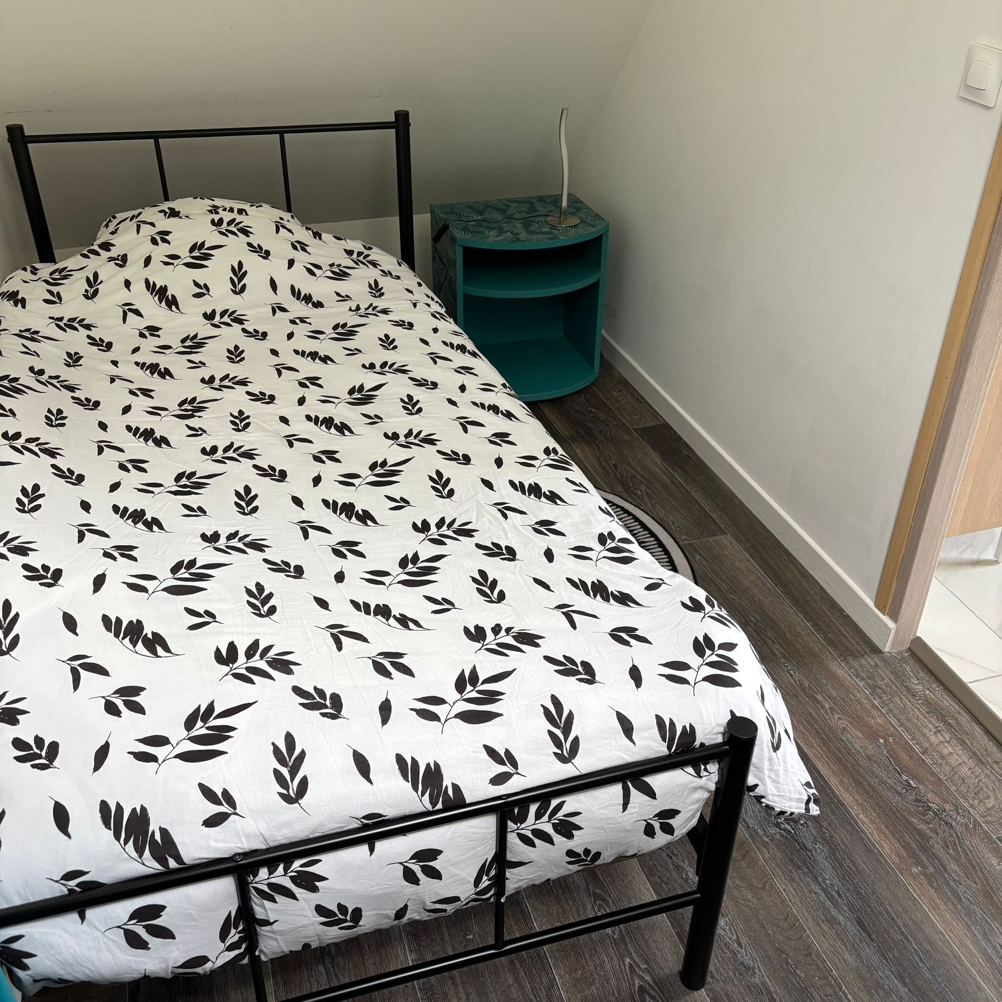 A bed with a leaf-patterned bedspread, a teal side table, and a white wall.
