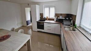 A compact kitchen with a dishwasher, microwave, and sink. There is a window providing natural light and a dining area with a table and chairs. - Quiet, fully-equipped house with garden (Maing)