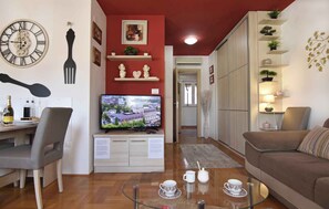 Een woonkamer met een televisie, een eethoek met een tafel en stoelen, een bank en decoratieve objecten op planken.