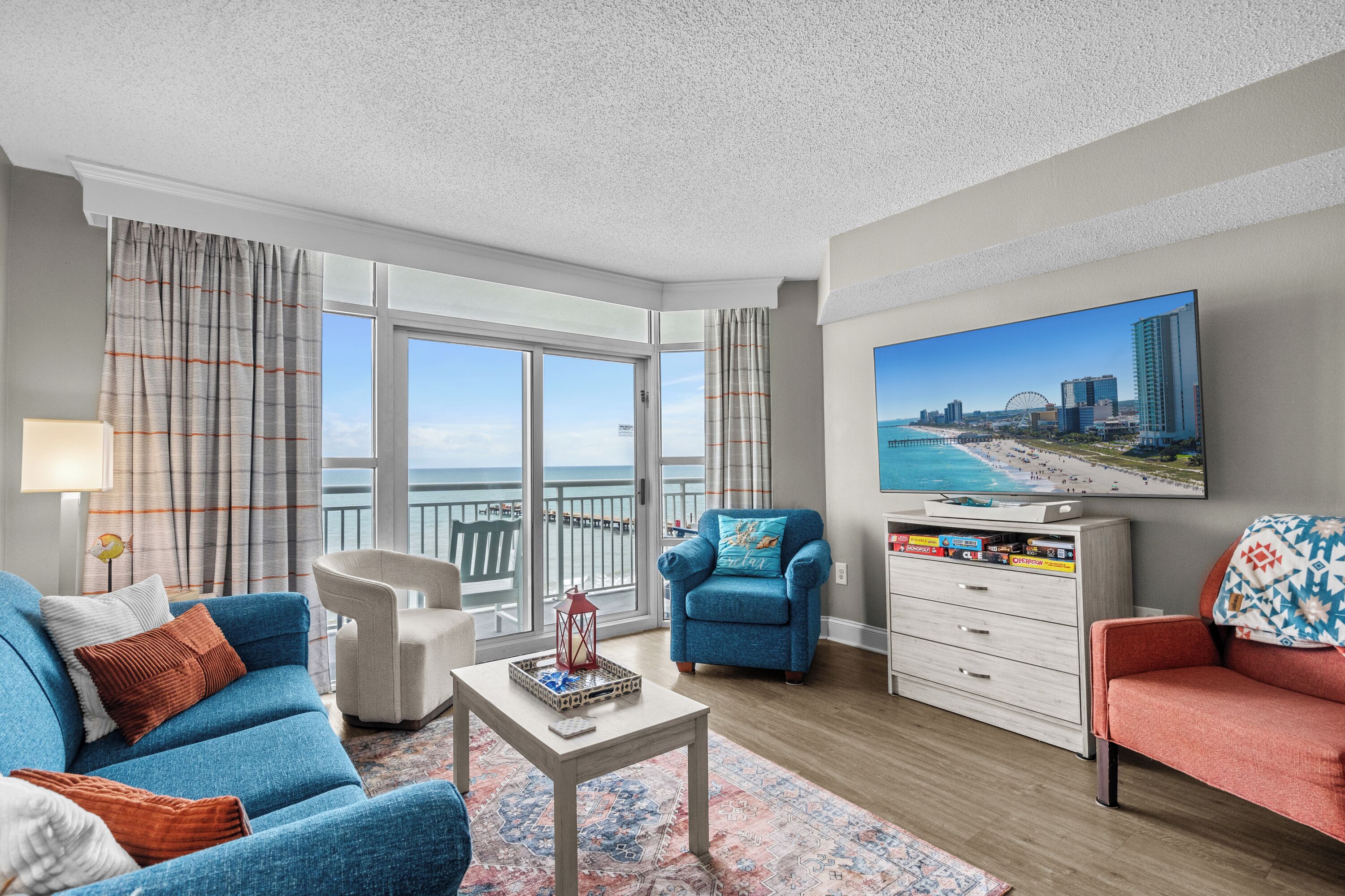 A modern living room with a large TV, comfortable seating, a view of the beach, and a balcony.