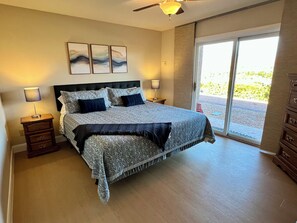 A bedroom with a bed, nightstands, a ceiling fan, artwork on the wall, and a sliding glass door leading to an outdoor area.