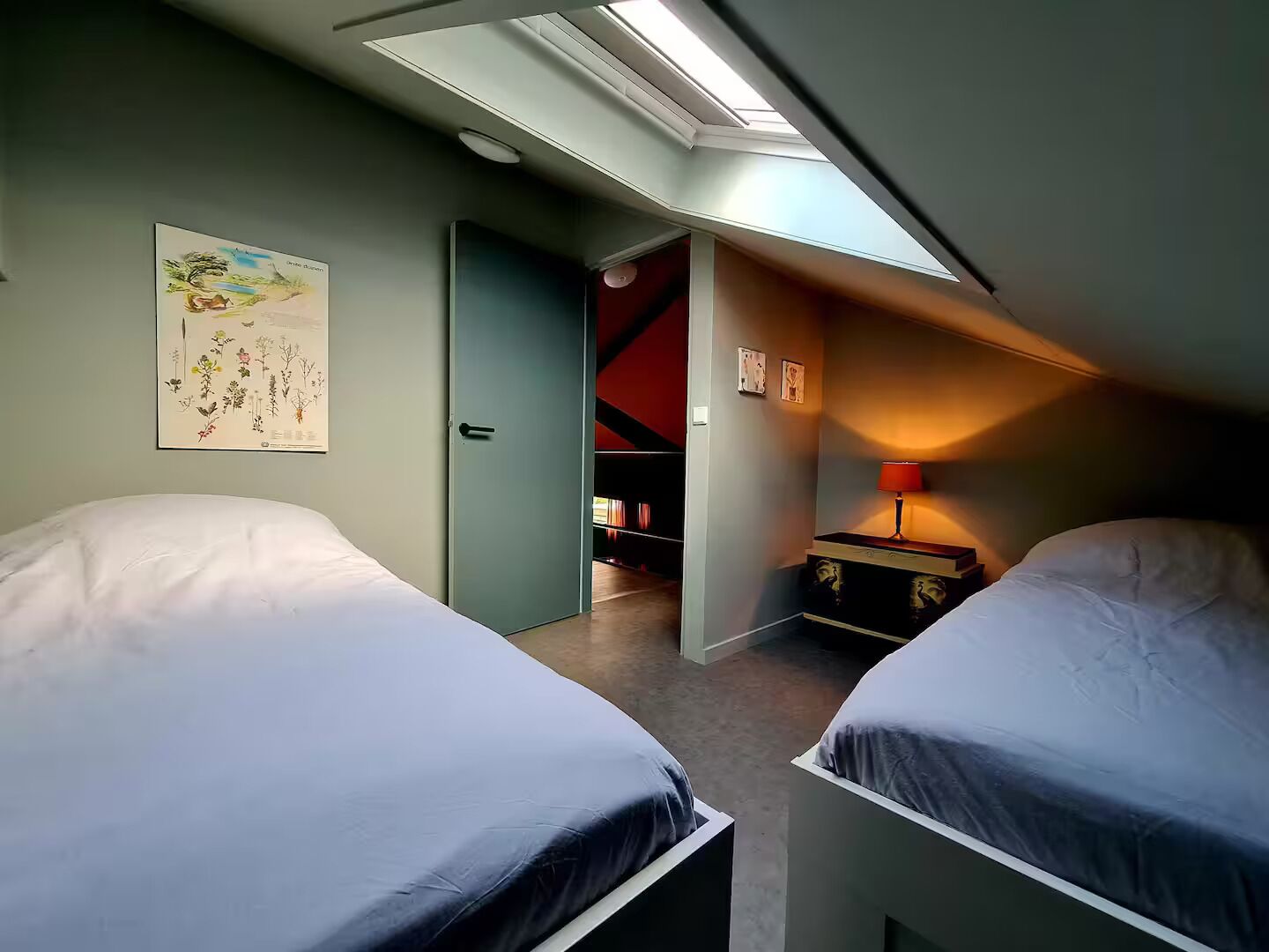 A bedroom with two beds, a skylight, a door leading to a hallway, and a small table with a lamp.