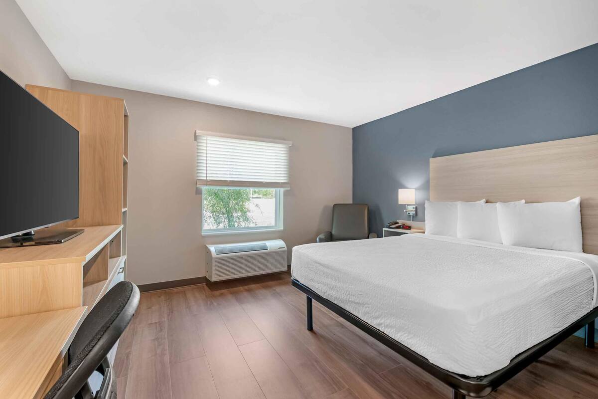 A hotel room with a bed, a desk with a TV, a chair, a window with blinds, and a nightstand with a lamp.