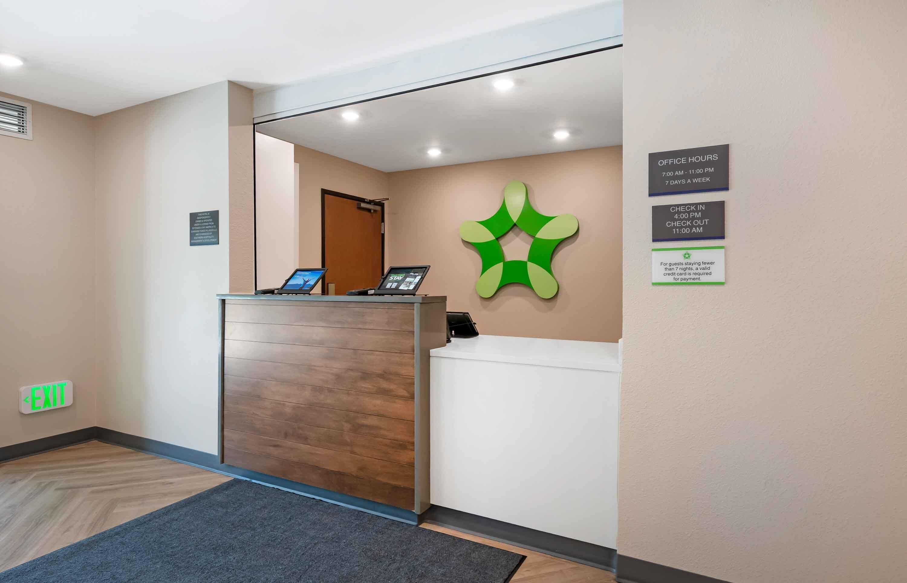 A hotel reception area with a wooden reception desk, a digital screen, and a green wall decoration. There are signs indicating office hours and check-in/check-out times.