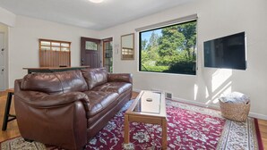 A living room with a brown leather sofa, a wooden coffee table, a red patterned rug, a large window with a view of trees, and a flat-screen TV mounted on the wall. - The Canon Hill Cottage (Spokane)