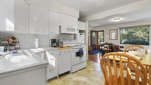 A modern kitchen with white cabinets, a microwave, an oven, and a coffee maker. There is a dining area with wooden chairs and a table. - The Canon Hill Cottage (Spokane)