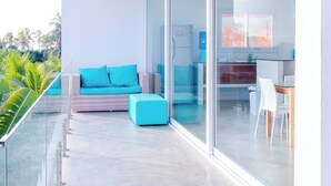 Un balcone moderno con ringhiera in vetro, un divano blu con cuscini e una zona cucina visibile attraverso le porte in vetro.