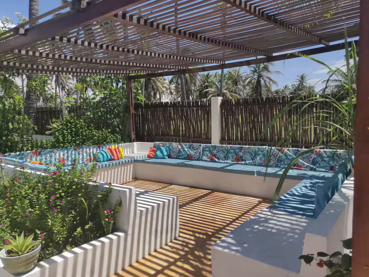 An outdoor seating area with a wooden bench and a cushioned sofa, surrounded by plants and a wooden fence.