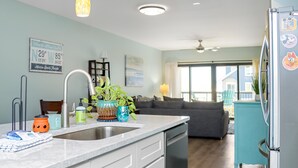 A kitchen with a sink, countertop, and cabinets. There is a refrigerator, a dining table, and a sofa in the background. - Club at Mexico Beach 1K Forgotten Cove (Port St. Joe)