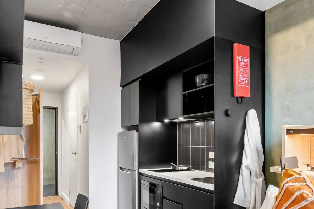A modern hotel room with a kitchenette, a bed with a white and orange blanket, and a small table with a chair.