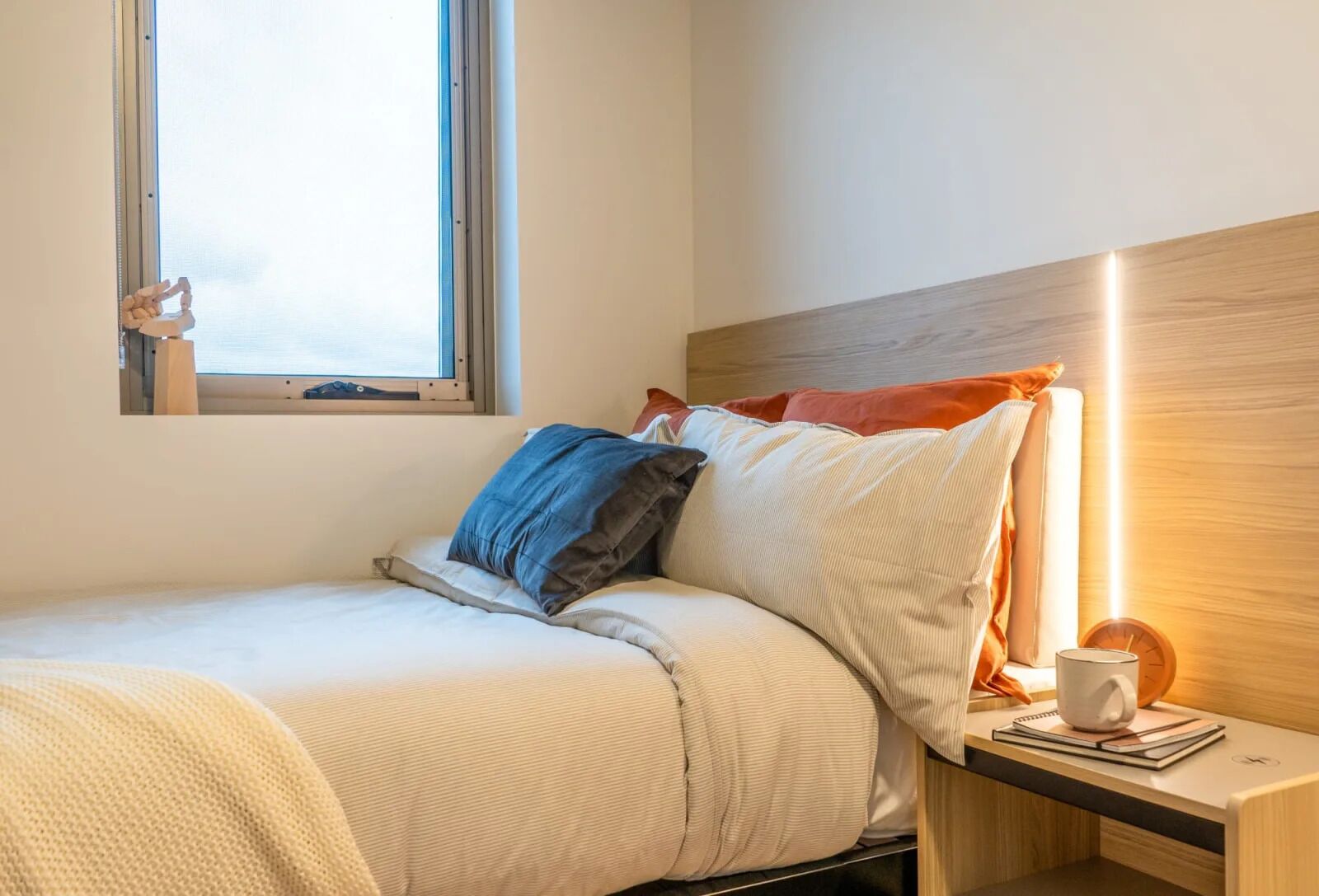A neatly made bed with a striped blanket, a bedside table with a lamp, and a window with a view of the sky.