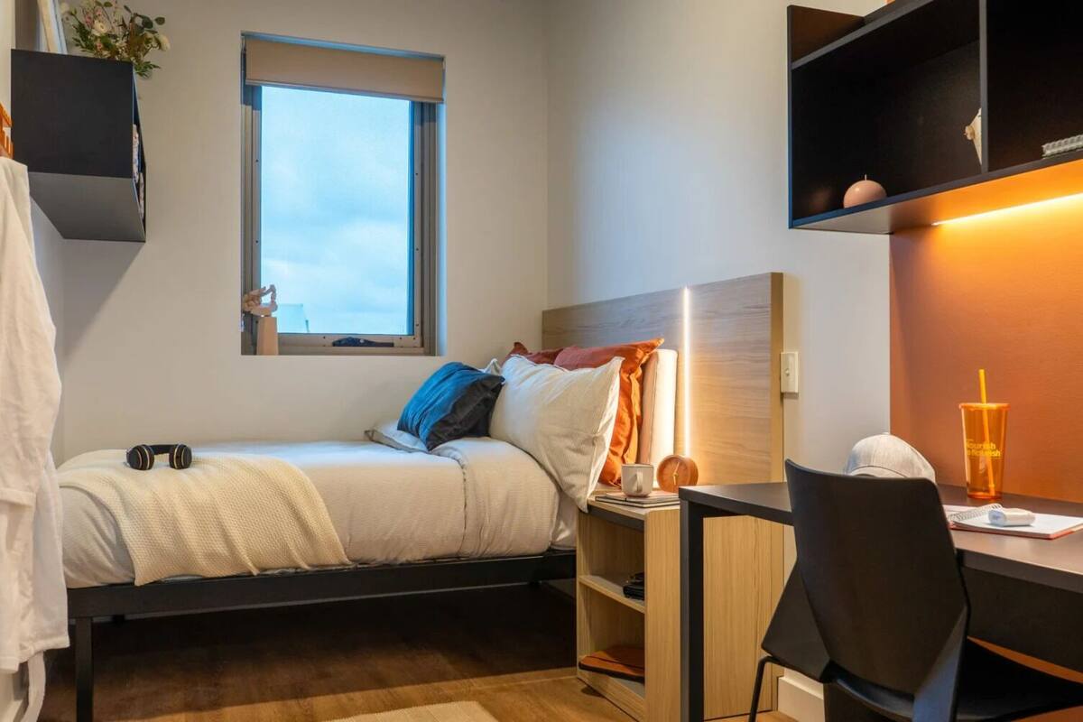 A compact hotel room with a bed, bedside table, chair, desk, and a window.