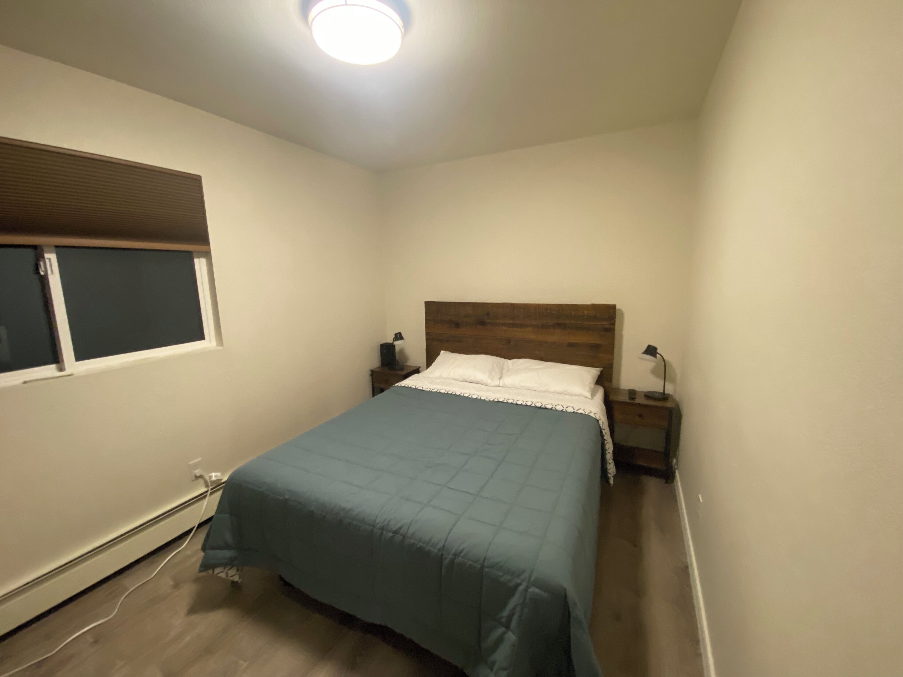 A bedroom with a bed, bedside tables, a window with blinds, and a ceiling light.