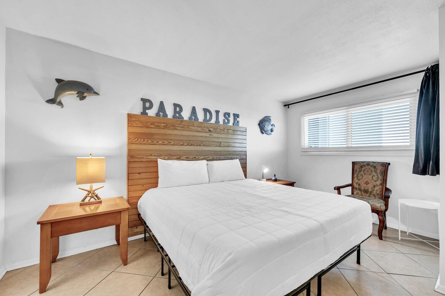 A bedroom with a large bed, a nightstand with a lamp, a chair, and a window with blinds.