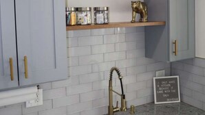 A kitchen with white tiled walls, a granite countertop, a stainless steel sink, and gold-colored fixtures. There are two shelves with decorative items and a framed picture above the sink. - Modern Cozy Apartment, Free Parking, Wi-Fi, Near Dining and Downtown. (Chicago)