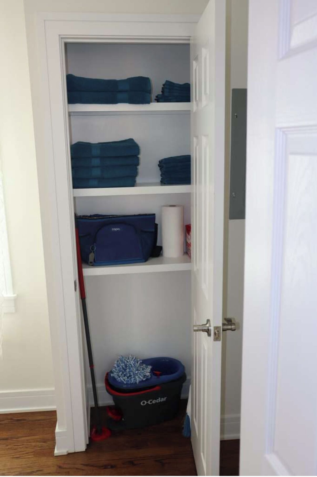 Ein Schrank mit Regalen, der gefaltete Kleidung, eine blaue Tasche und einen Wischmopp beinhaltet.