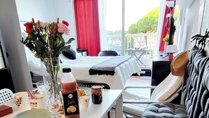 A cozy room with a dining table set with a bottle, glasses, and a plate. There is a sofa with a colorful blanket and a hat on it. A window with red curtains is visible, and a balcony with a table and chairs is seen in the background.