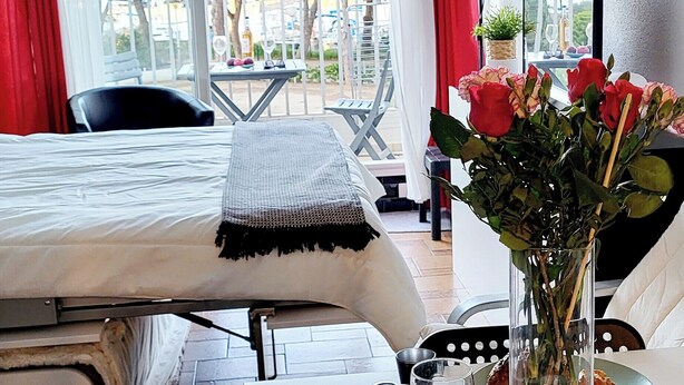 A hotel room with a bed, a table with a vase of flowers, a chair, and a balcony with a view of trees and a table outside.