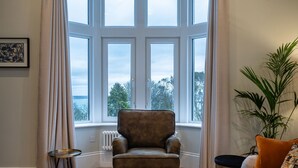 Une pièce dotée d’une grande baie vitrée, d’un fauteuil marron, d’une petite table ronde, d’un canapé avec des coussins et d’une plante en pot.