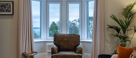 A room with a large bay window, a brown armchair, a round side table, a sofa with cushions, and a potted plant.