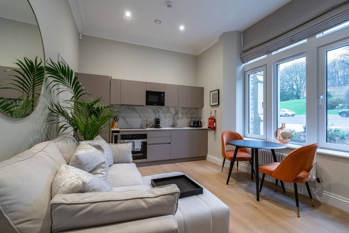 A modern living room with a sofa, a dining table with chairs, a kitchen area with cabinets and appliances, and a large window with a view of the outside.
