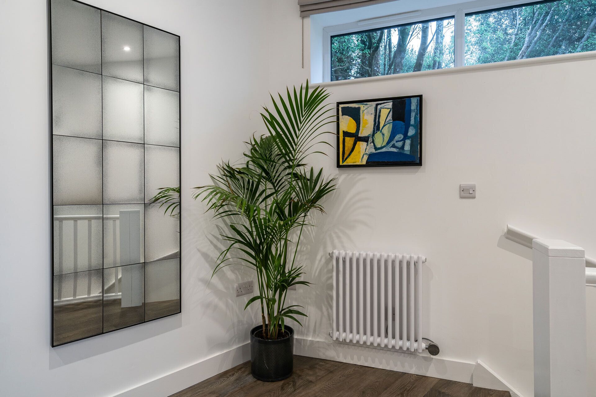 A modern interior with a large mirror, a potted plant, a radiator, and a window with a view of trees.