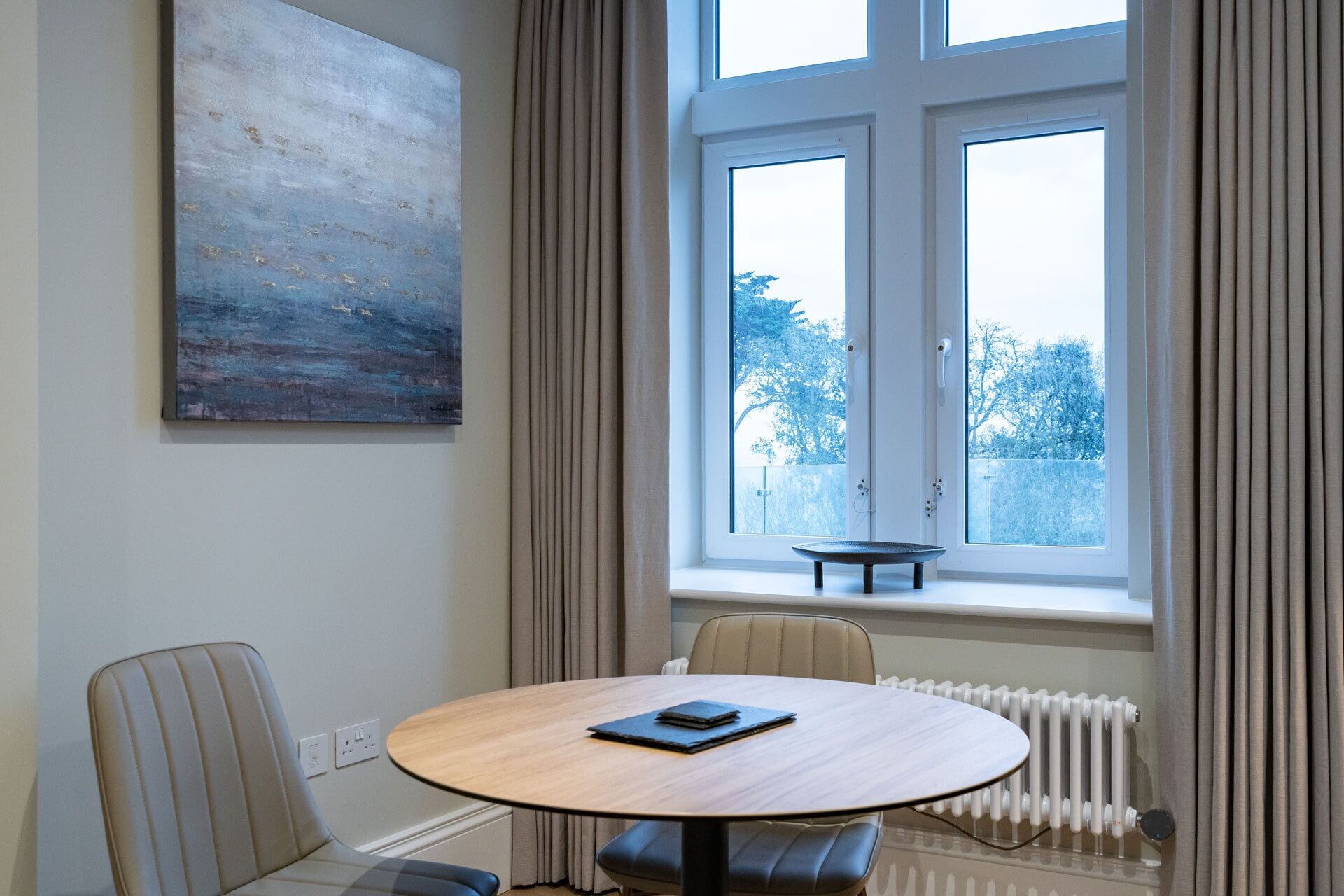 Ein Zimmer mit einem runden Tisch, zwei Stühlen, einer Heizung und einem Fenster mit Vorhängen.