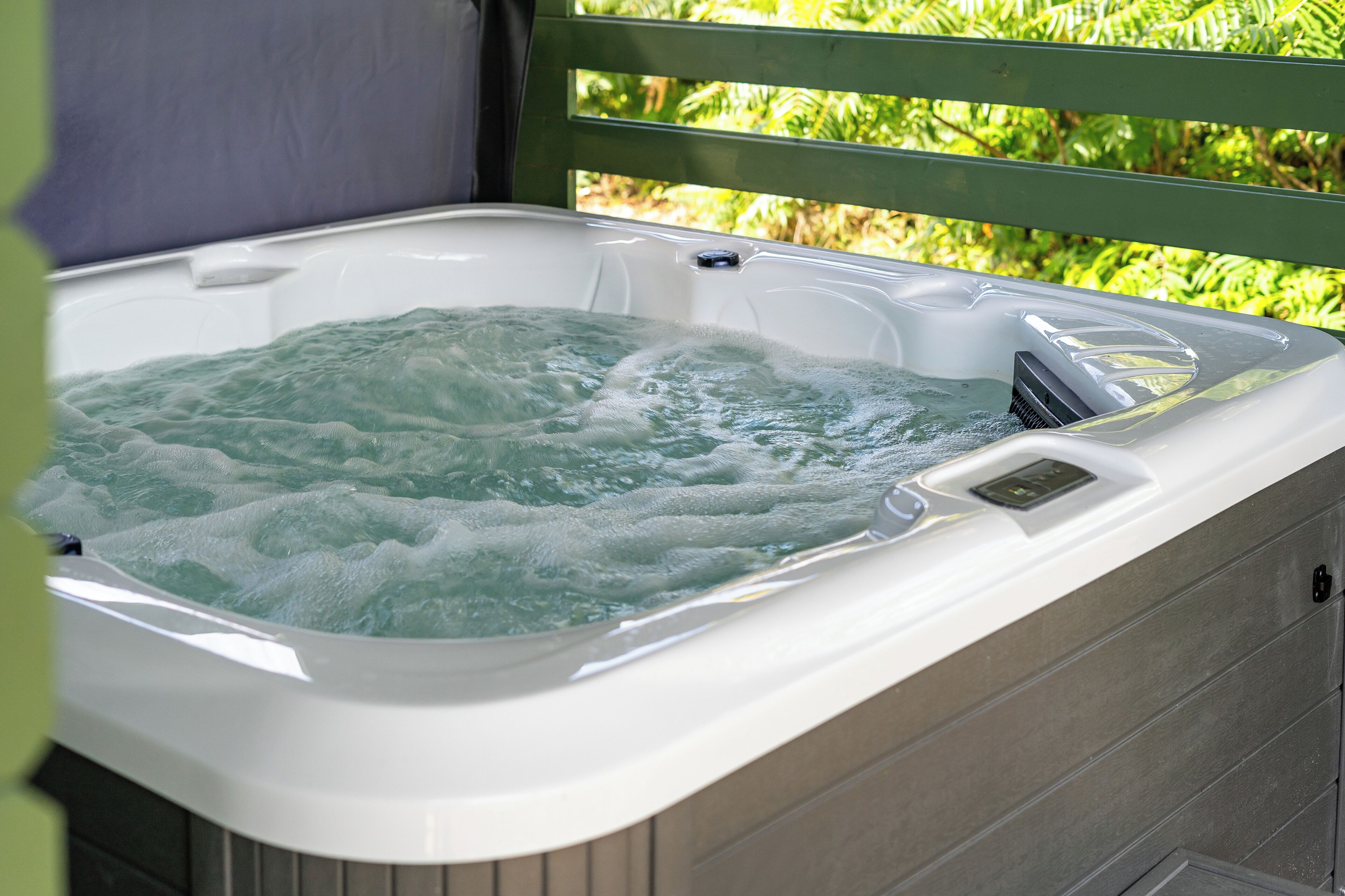 A hot tub with a control panel, surrounded by a wooden deck and greenery.
