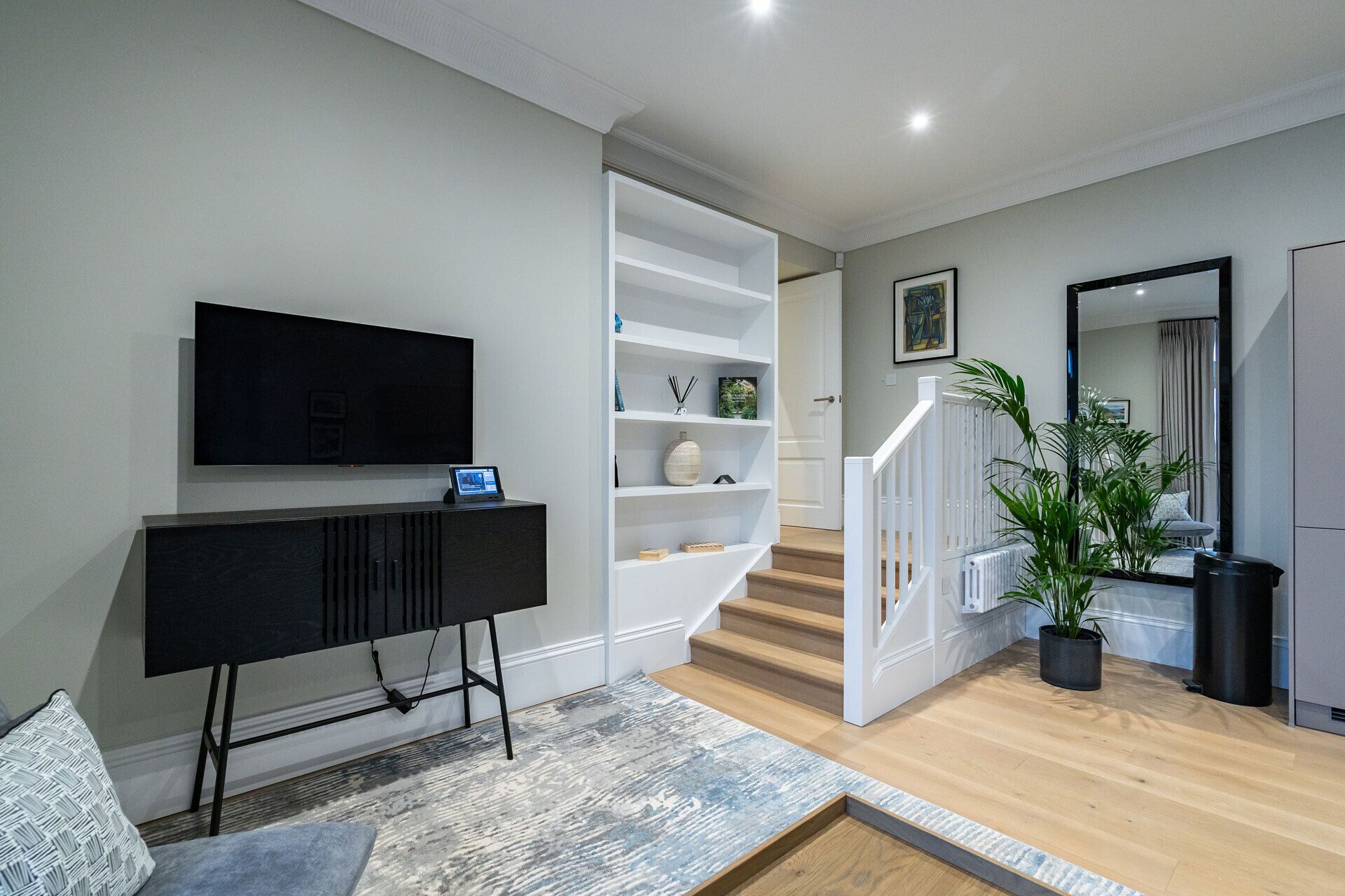 A modern living room with a flat-screen TV, a black entertainment unit, a white shelving unit, a staircase, and a large mirror.